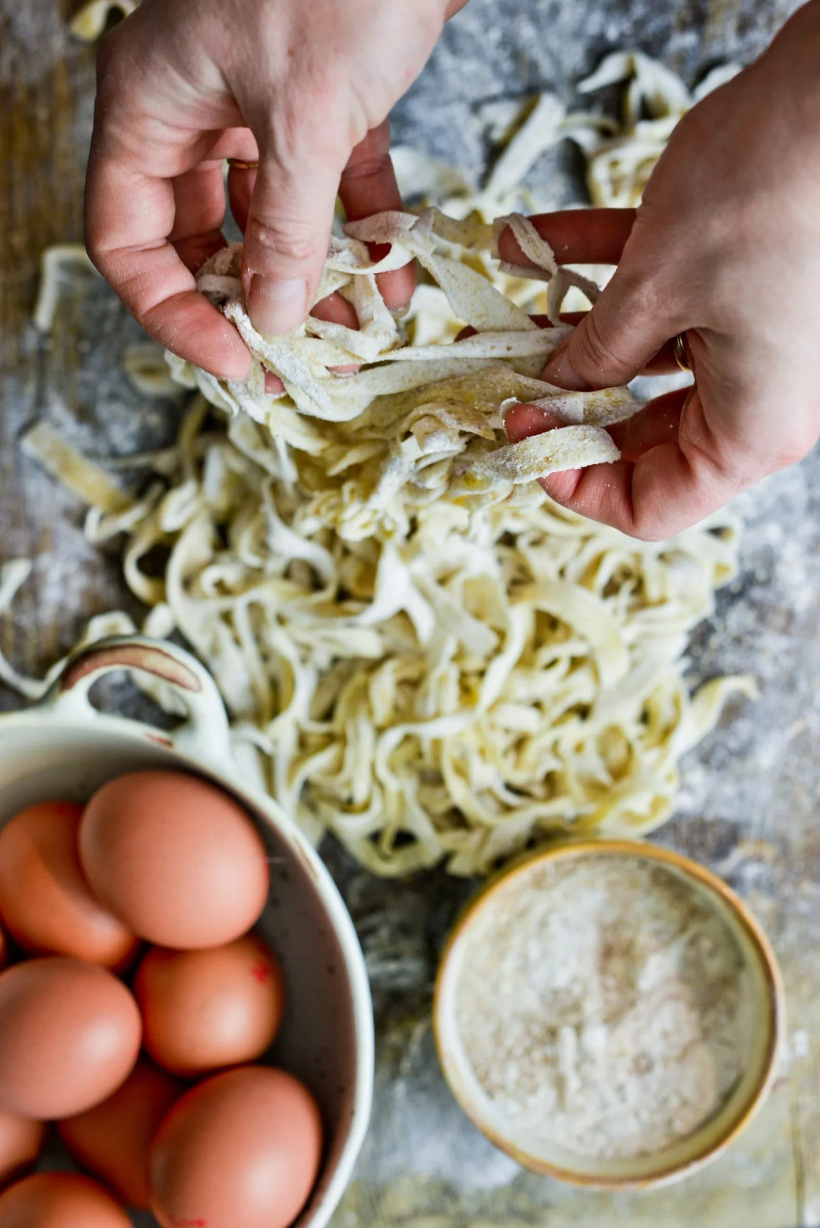 Hands laying out fresh tagliatelle next to the eggs and flour